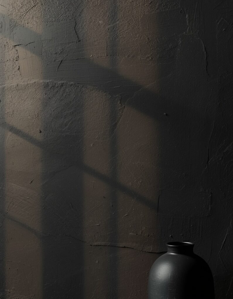 A deep charcoal limewash wall surface photographed as an architectural macro showing its characteristic mineral mottling — each section catching afternoon raking light differently, the surface appearing to glow from within, tonal variation from darker to slightly lighter areas visible across the plaster surface, one matte black ceramic vessel partially visible at lower right