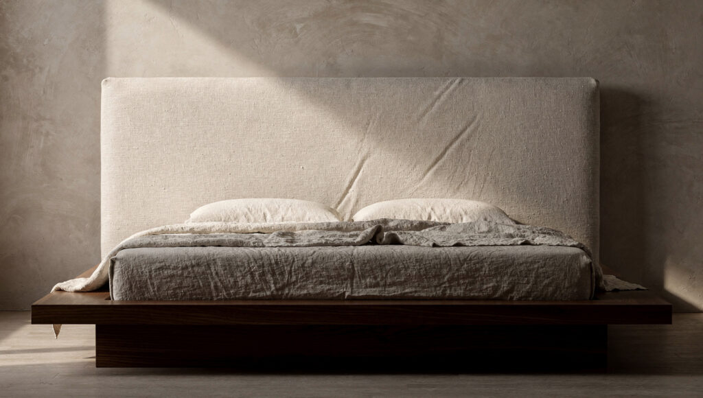 A low dark walnut platform bed with a single upholstered headboard panel in heavyweight stonewashed natural oat linen showing visible natural crease and texture, the walnut frame visible at the base and edges creating contrast between wood and textile, with matching oat linen bedding
