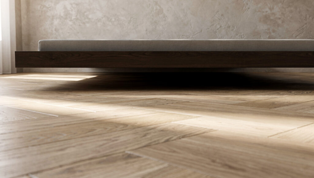 A low-angle view showing light oak herringbone flooring running completely unbroken beneath the edge of a dark walnut platform bed with no box spring, bed skirt, gap, or storage visible — the floor plane continuous from the wall to beneath the bed frame