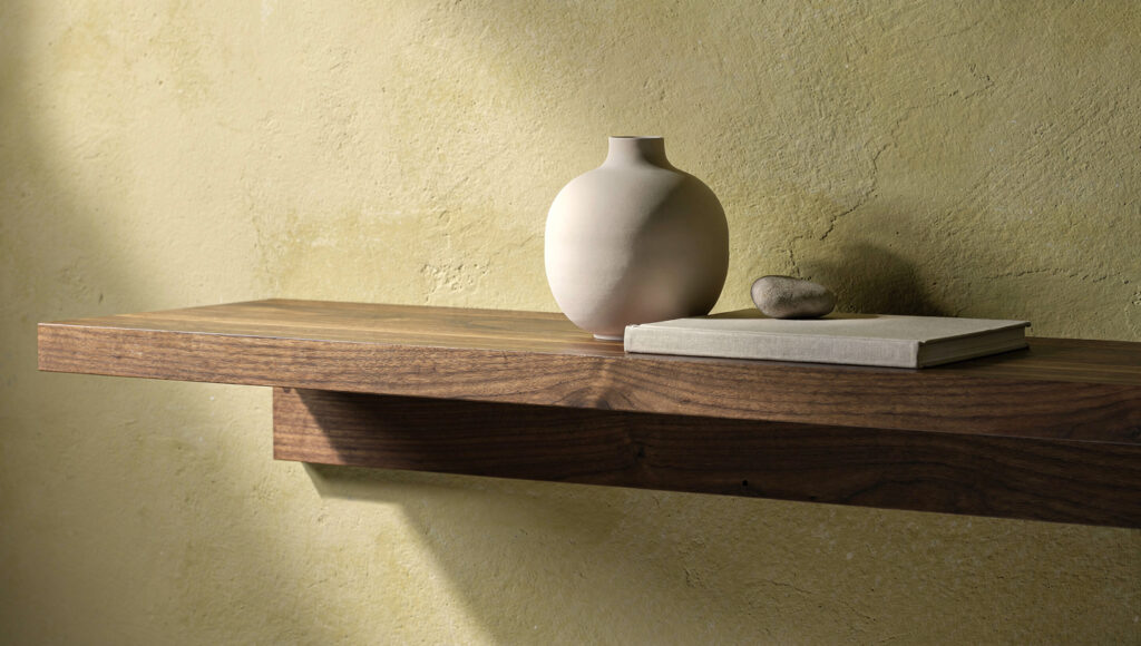 A solid walnut floating shelf 1.75 inches thick with a concealed bracket, styled with three objects — a ceramic vessel, a flat book, a stone — spaced unevenly with clear shelf on either side