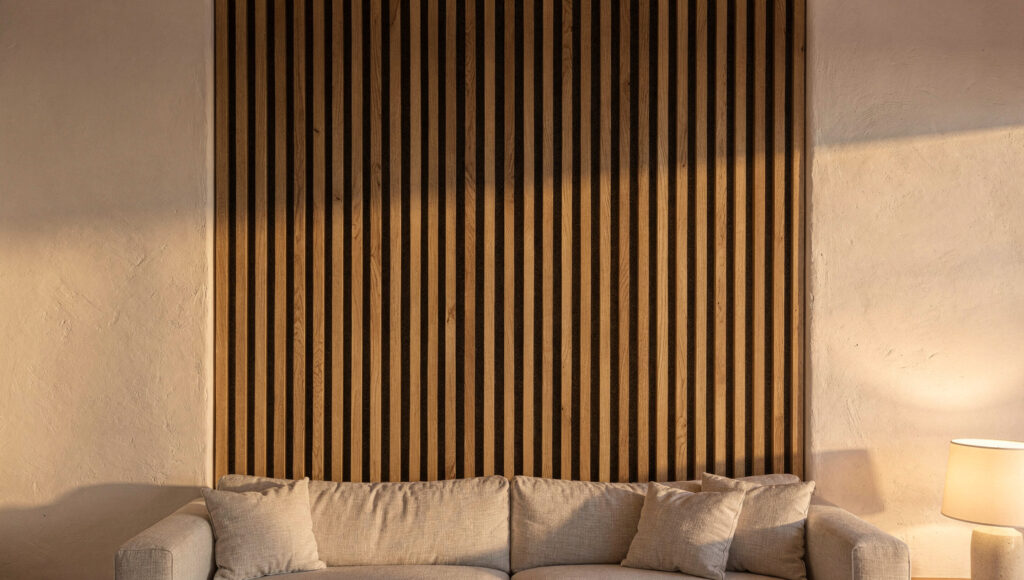 Vertical white oak acoustic slat panels as a defined zone behind a low linen sofa, dark felt visible in the shadow gaps, plain plaster on either side, under warm raking side light