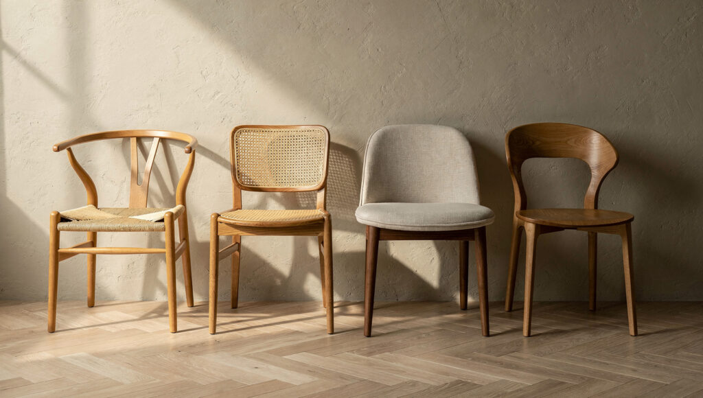 Four Japandi dining chair forms displayed side by side against a warm greige limewash wall — a solid ash wishbone chair with paper cord seat, a natural cane back oak chair, an oat linen upholstered walnut frame chair, and a solid oak sculptural chair — all in natural materials with matte finishes on herringbone oak flooring
