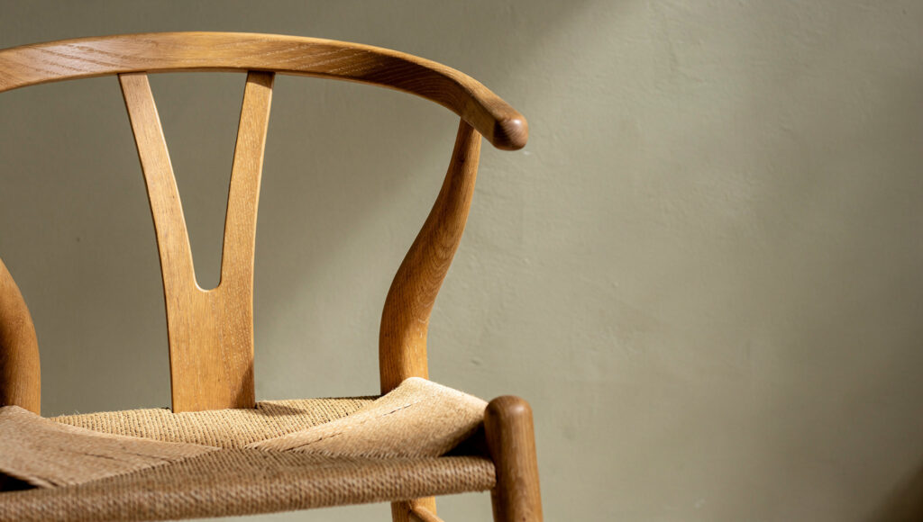 A solid natural ash wishbone dining chair against a warm greige limewash wall showing the Y-form back joint in detail — the honest wood-to-wood joinery visible at the junction, the woven paper cord seat beneath, and the warm greige wall clearly visible through the negative space of the Y-back