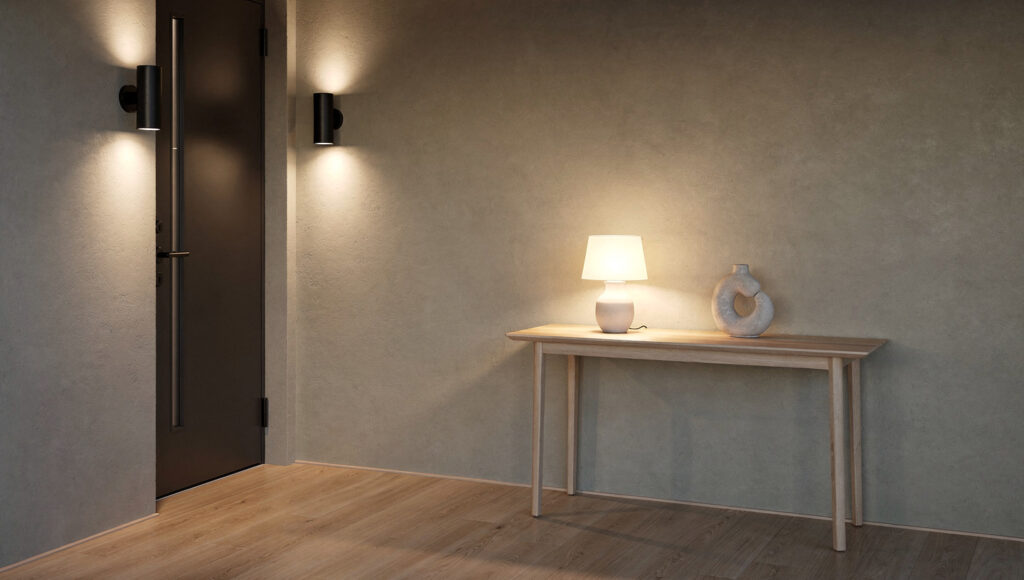 A Japandi entryway at dusk showing two warm light sources — a matte black wall sconce beside the front door and a small ceramic table lamp on the console, both glowing at 2700K with no overhead light active, creating layered warm illumination against textured greige limewash walls