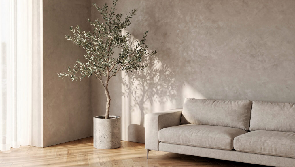 A 6-foot faux olive tree in a raw concrete cylinder planter placed in the corner where two warm greige limewash walls meet in a Japandi living room, morning side light creating organic leaf shadow patterns on the adjacent wall surface, the asymmetric canopy extending into the room with a linen sofa partially visible to the right