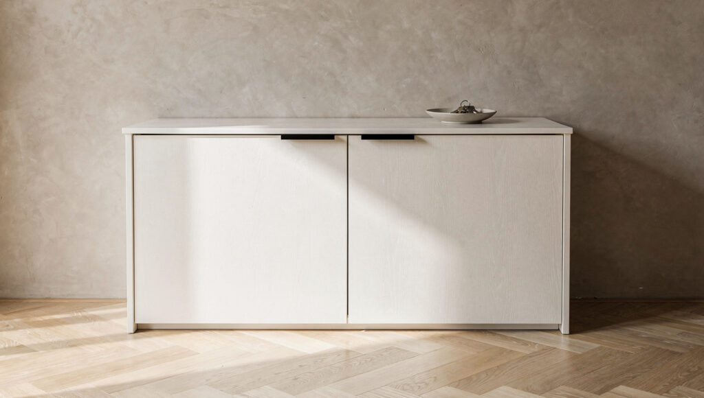 A closed white oak shoe cabinet in getabako style with flat-panel doors and slim recessed matte black pulls completely concealing all footwear, the floor in front completely clear, one ceramic dish with keys on top against a warm greige limewash wall
