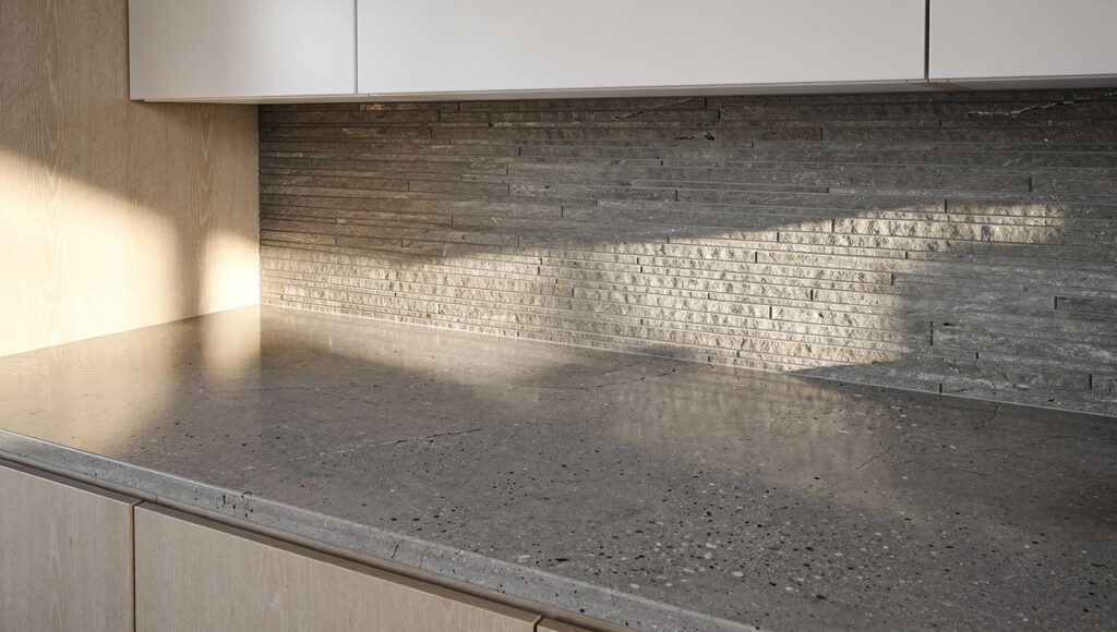 A Japandi kitchen countertop and backsplash detail showing a honed concrete-effect quartz countertop in warm gray with matte mineral texture absorbing morning light, paired with a warm gray natural stone backsplash in slim stacked horizontal tiles, white oak lower cabinet edge visible left and upper cabinet base visible above