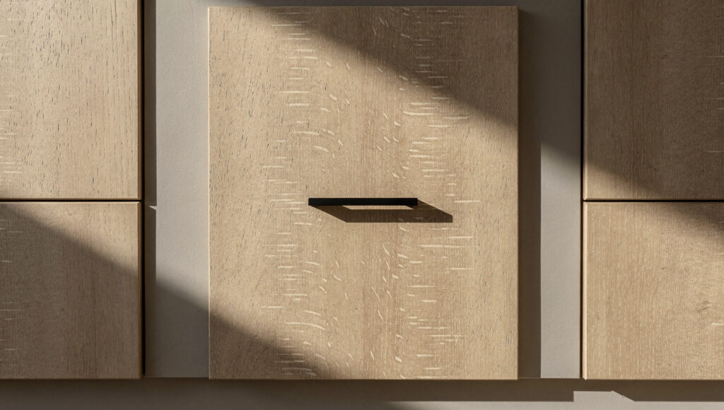 A rift-sawn white oak flat-panel cabinet door showing straight vertical grain lines running consistently from top to bottom with medullary ray fleck pattern visible, a slim 4-inch matte black square bar pull at center, matte wood surface absorbing morning light, adjacent doors partially visible