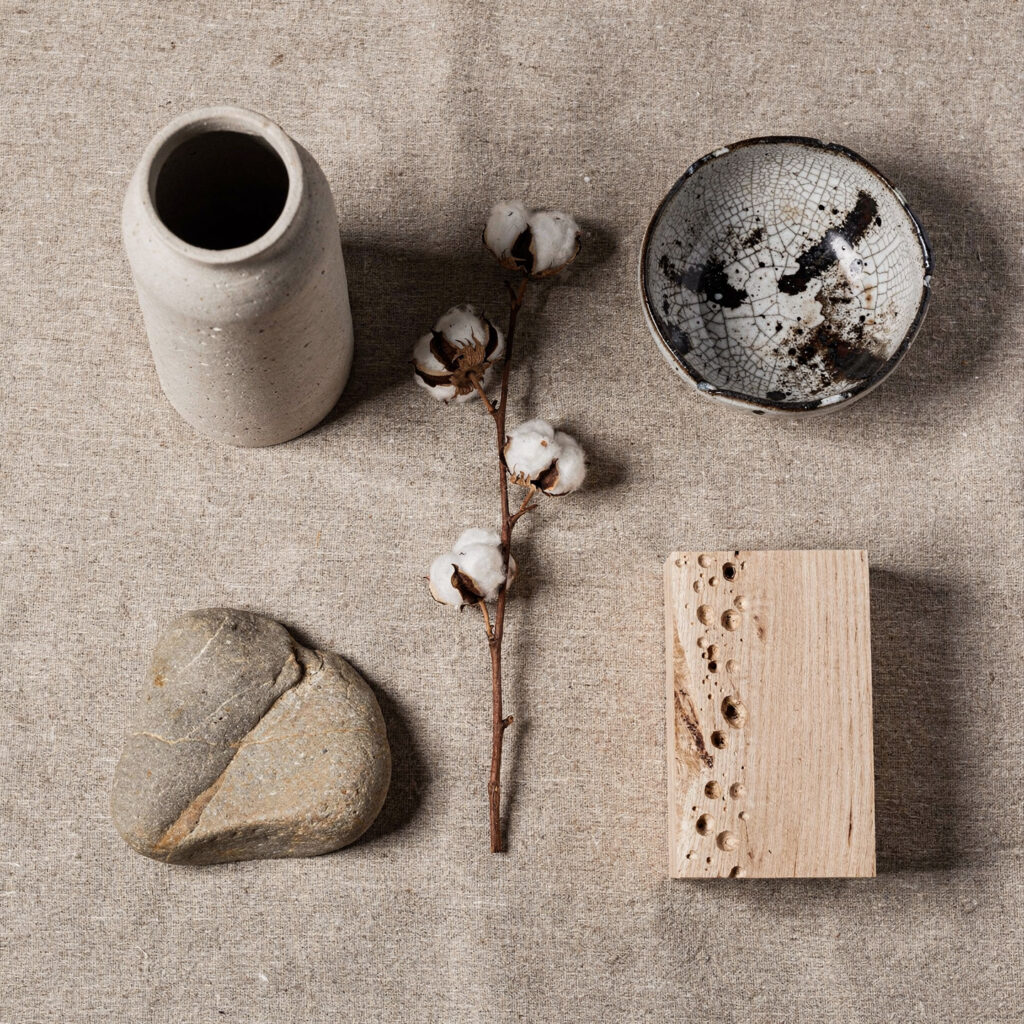 A flat lay on raw linen showing the five permitted Japandi shelf object types — a matte unglazed stone ceramic vessel, a raku crackle-glaze bowl with smoke marks, a dried cotton stem, a smooth river stone with natural mineral variation, and a small piece of unfinished white oak — all matte, all natural