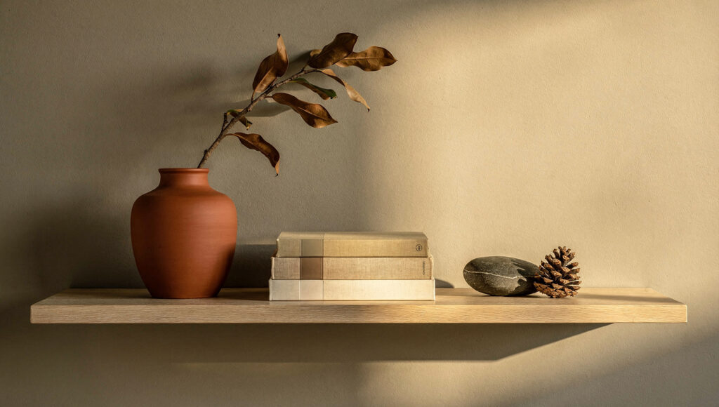 A white oak floating shelf styled for autumn showing a wide matte terracotta ceramic vessel with a dried magnolia branch as the anchor, four horizontally stacked warm oat and sand toned books in the center, and one smooth dark river stone with a pine cone in the release zone with clear space beyond