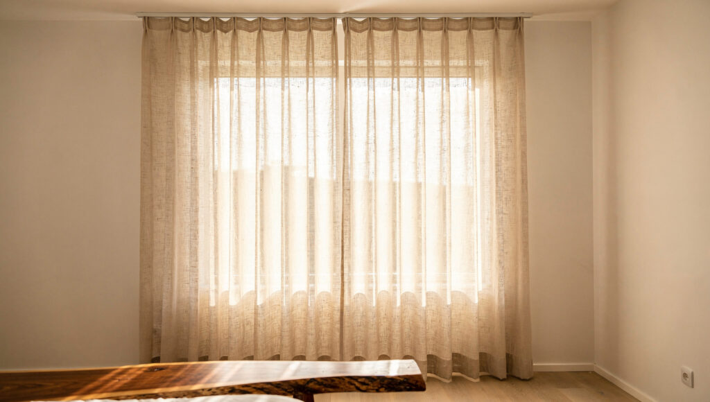A bedroom window with sheer undyed linen curtains showing the natural weave texture against bright daylight passing through, the fabric diffusing the light into warm even illumination rather than harsh direct sun, a live-edge wood bed frame corner visible in the foreground