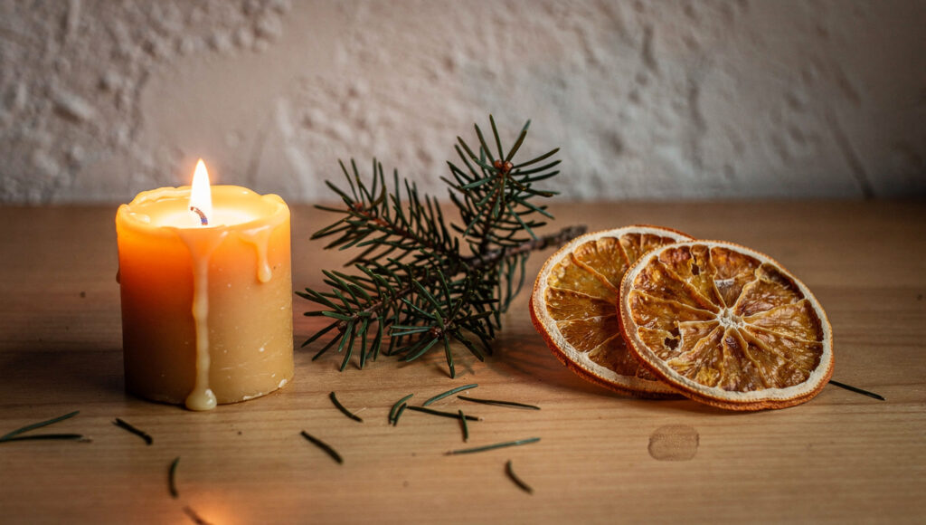 Three objects spaced widely on a raw oak surface — a burning beeswax candle, a sprig of fresh fir, and two dried orange slices — lit mainly by the candle flame