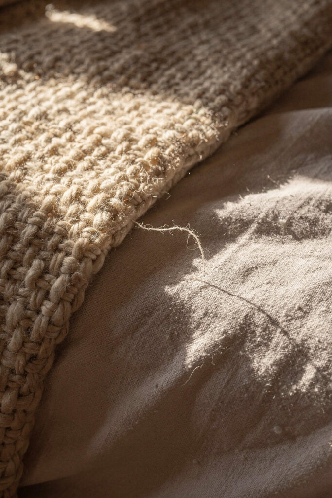 A coarse cream wool throw folded over smooth washed linen bedding, the two textures meeting under low raking autumn light