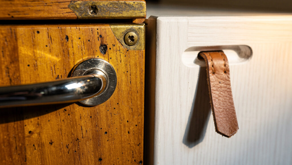 A teak drawer front with a chrome pull and brass fixing beside a white oak drawer front with a routed finger notch and a worn leather tab.