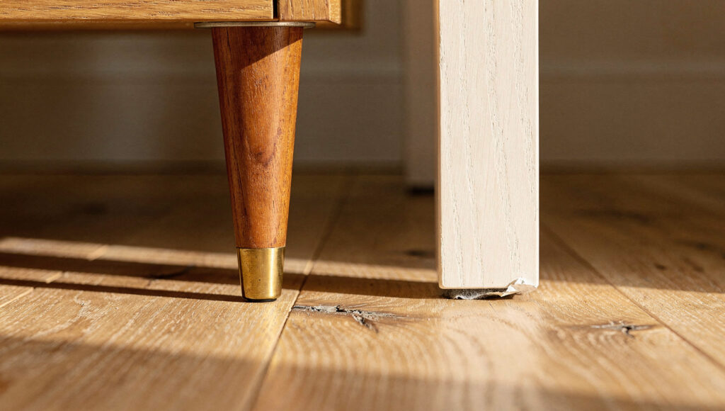 A close floor-level view of a splayed brass-tipped teak leg beside a straight square oak leg, showing where each foot lands relative to the piece above.