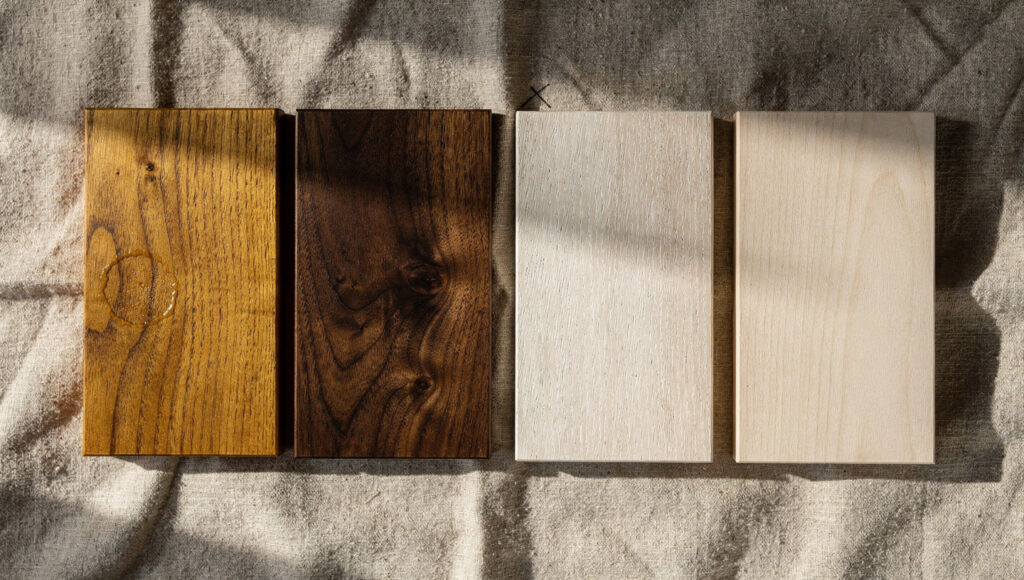 Four timber samples laid on oatmeal linen in raking light, gold-brown teak and dark walnut beside pale wire-brushed white oak and ash.